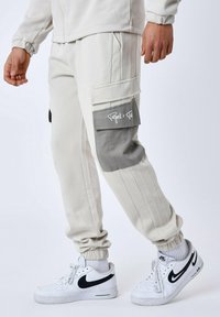 Lichtgrijze joggerbroeken met elastische boorden, voorzien van twee zijcargo pockets, waarvan één met een donkergrijze flap, en witte sneakers.