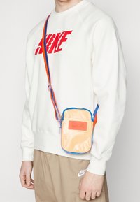 Persona que lleva una camisa blanca de manga larga con texto rojo, pantalones beige de Nike y una pequeña bolsa crossbody beige con una correa azul y naranja.