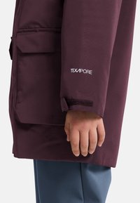 Veste imperméable bordeaux avec marquage Texapore, dotée d'une poche à rabat et de détails au niveau des poignets en Velcro, portée avec un pantalon bleu clair.