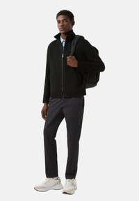 Sort fleecejakke med høj krave, grå bukser og hvide sneakers. Personen bærer en sort rygsæk. Enkel design.