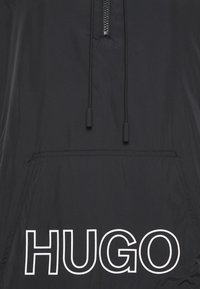 Mustaa kevyttä anorakkia, jossa on puolivetoketju, kiristettävä huppu ja etupuolella kengurupusseja. "HUGO"-logo painettuna valkoisena etupuolelle.