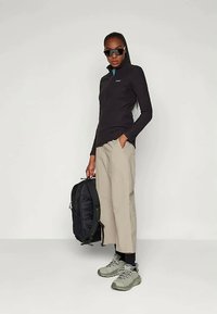 Fekete hosszú ujjú felsőt és bézs színű, széles lábú nadrágot viselő nő, fekete hátizsákot tart, napszemüveget visel, és szürke túracipő van rajta.