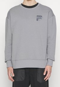 Šedá mikina vyrobená z jemné bavlněné směsi, s kulatým výstřihem, dlouhými rukávy, ribbovanými manžetami a jemným potiskem loga na hrudi.