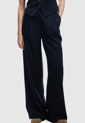 Person, die eine marineblaue, gestreifte, weite Hose mit seitlichen Taschen und braunen Schuhen trägt, vor einem schlichten Hintergrund.