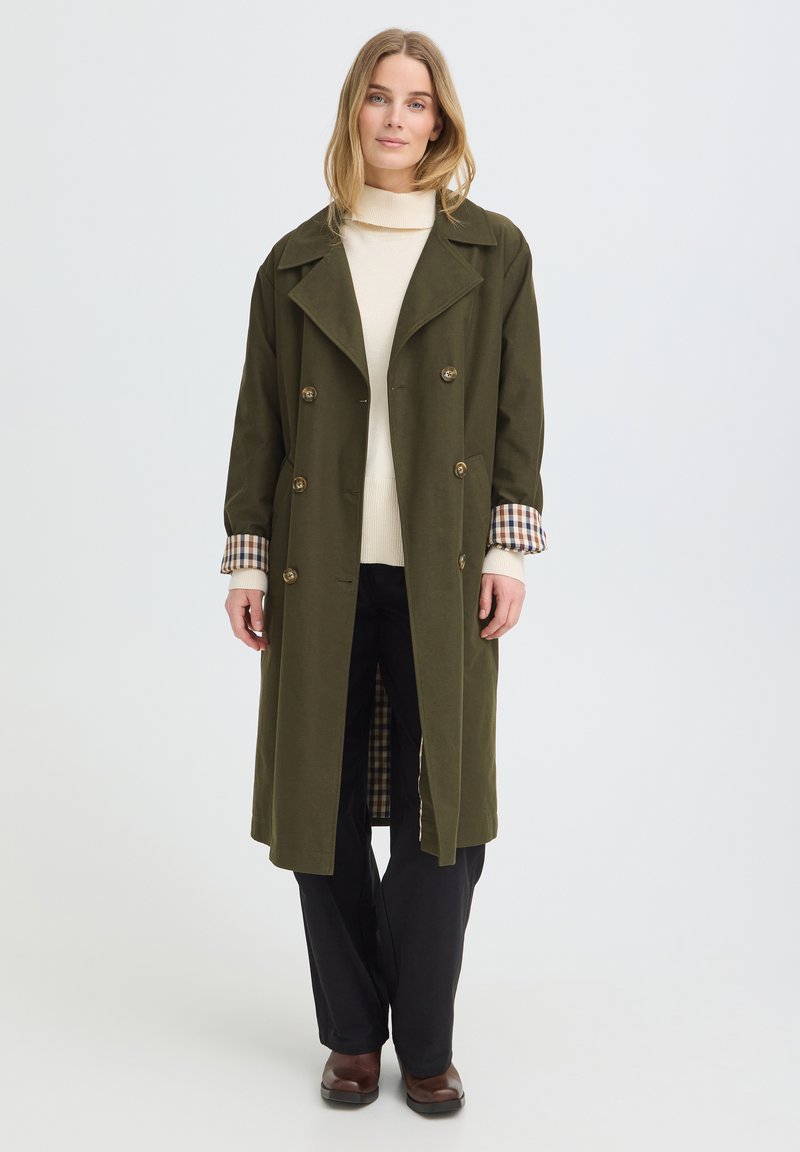 Femme portant un manteau croisé vert olive avec des poignets à carreaux, un pull à col roulé crème, un pantalon noir et des chaussures marron, debout sur un fond uni.