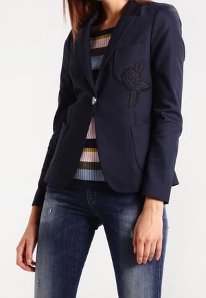 Kvinde iført en navy blazer med et blomsterpatch, en stribet sweater og slidte blå jeans, stående med afslappede arme.