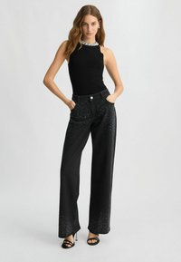 Top senza maniche nero con scollatura di gioielli, abbinato a jeans neri a vita alta con dettagli di strass sulle gambe inferiori.