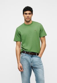 Άνδρας που φοράει πράσινη κοντομάνικη μπλούζα με μικρό λογότυπο στο στήθος, ανοιχτο-μπλε τζιν και καφέ υφαντή ζώνη, όρθιος.