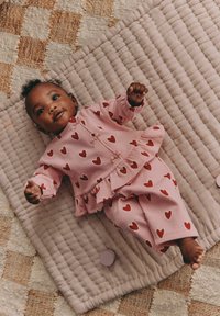 Rosa tvådelad babyset med röda hjärtmönster, bestående av en jacka med volanger och matchande byxor, utlagt på en texturerad, blekrosa matta.