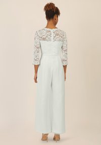 Weißer Jumpsuit mit Spitzenoberteil und Dreiviertelärmeln, mit einem Reißverschluss auf der Rückseite und weitem Hosenbein.