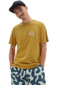 Camiseta de manga corta color mostaza con logo impreso en blanco, combinada con pantalones cortos de camuflaje en tonos azul y azul claro. Diseño simple y casual.