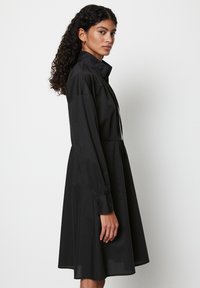 Schwarzes Kleid mit Kragen, langen Ärmeln, elastischen Bündchen und einem Taillengürtel. Es hat einen ausgestellten Rock und eine glatte Textur.