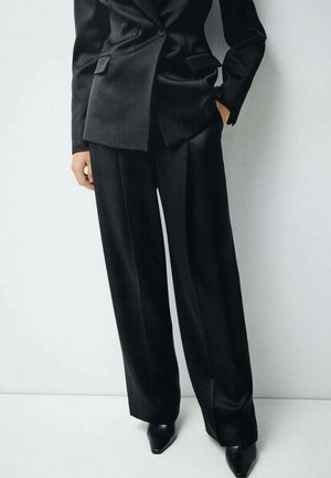 Costume noir ajusté avec un blazer croisé et un pantalon à jambes larges. Tissu lisse et brillant, avec une coupe structurée et des revers accentués.
