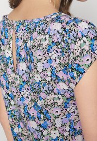 Blusa floreale con un motivo nero, verde, blu e viola. Presenta un collo rotondo e un dettaglio a bottoni sul retro, con maniche corte.