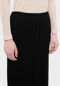 Sorte strikket skjørt med en teksturert finish, med en bred midjebånd og sømdetaljer. Modellen har hånden avslappet på siden av skjørtet.
