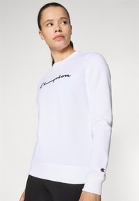 Sweatshirt blanc avec un logo "Champion" noir en cursive sur la poitrine. Manches longues, poignets et ourlet côtelés. Le tissu a une texture lisse.