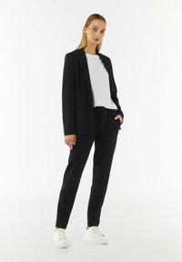 Zwarte blazer met een getailleerde pasvorm over een wit T-shirt; gecombineerd met zwarte tapered broeken en witte sneakers. Gladde, hoogwaardige stof.