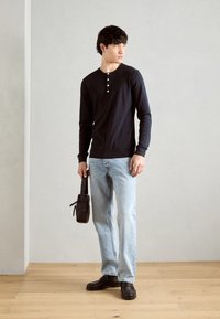 Marineblå langermet henley-skjorte med tre knapper, lyseblå rette jeans, svart veske og svarte skinnsko på tregulv.
