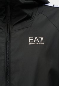 Μαύρο μπουφάν με φερμουάρ μπροστά και λευκό λογότυπο EA7 Emporio Armani στο στήθος, με μια μικρή λευκή λωρίδα υφάσματος κοντά στον ώμο.