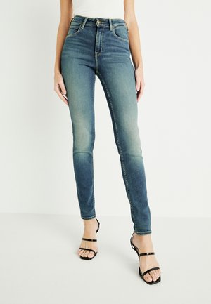 Jeansy skinny w kolorze niebieskim z wysokim stanem, wykonane z lekko przetartego denimu o gładkiej fakturze, z przednimi kieszeniami i zapięciem na guzik, w połączeniu z czarnymi sandałami na paski.