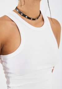 White ribbed tank top with a high neckline and wide armholes, paired with a black choker featuring silver studs.