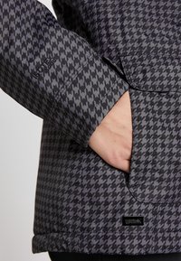 Main dans la poche d'une veste noire et grise à motif pied-de-poule avec logos de la marque visibles sur la manche et près de la poche.