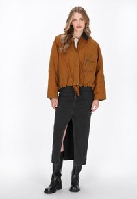 Bruine, cropped jas met een voorzak, zwarte kraag en een trekkoord aan de onderzijde; gecombineerd met een zwarte, enkellange rok met een vooropening en laarzen.