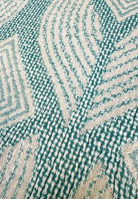 Geweven textiel met geometrische patronen in teal en crème. De textuur is tastbaar met een verhoogd, geweven oppervlak en ingewikkelde details.