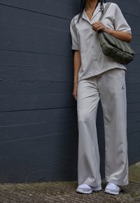 Personne portant une chemise à manches courtes boutonnée gris clair assortie à un pantalon avec un petit logo Jordan noir, tenant un sac d'épaule matelassé vert olive.