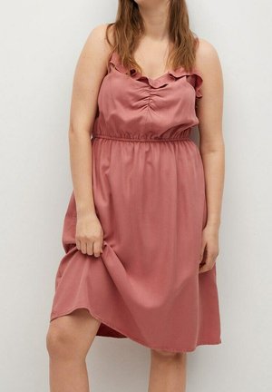 Femme portant une robe sans manches d'une longueur au genou de couleur rose poussiéreuse avec un col à volants, tenant l'ourlet d'une main sur un fond clair uni.