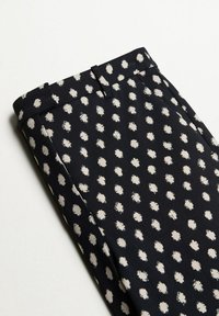 Pantalon noir à pois blancs irréguliers, avec passants de ceinture et poches latérales, plié soigneusement sur un fond blanc.