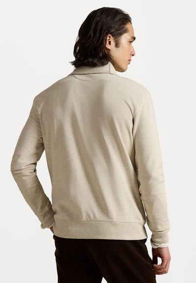Beige pletený svetr s dlouhým rukávem a stojáčkovým límcem, jemná textura, žebrované manžety a lem, nošený osobou otočenou zády.