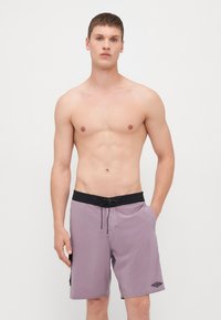 Lilla svømmeshorts med svart midje, snøring, sidelommer, og en liten logopatch på venstre ben.