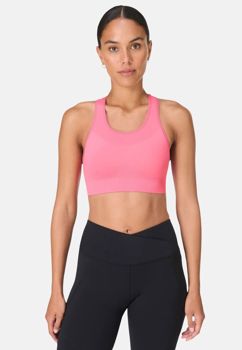 Femme aux cheveux foncés portant un soutien-gorge de sport rose et un legging noir, debout devant un fond clair uni, face à l'avant.