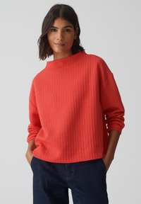 Pull en corail côtelé avec un col haut, coupe décontractée et manches longues. Assorti à un jean bleu foncé, affichant un style décontracté.