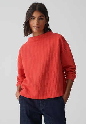 Maglione a coste color corallo con collo alto, vestibilità rilassata e maniche lunghe. Abbinato a jeans blu scuro, che mostrano uno stile casual.