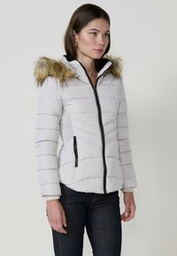 Világosszürke puffertakaró, bunda díszítéssel ellátott kapucnival, cipzáras záródással és oldalsó zsebekkel. Különlegessége a tűzött textúra és a fekete cipzár díszítések.