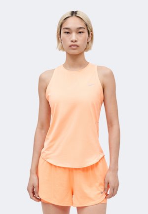 Femme portant un haut de sport sans manches et un short assortis couleur pêche avec logos Nike, debout devant un fond clair uni.