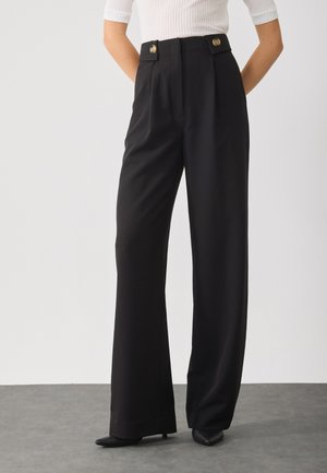 Woman wearing high-waisted black wide-leg trousers with buttoned waistband and pointed black shoes, standing on gray floor.