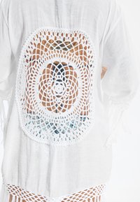 Copricostume bianco leggero con un grande motivo all'uncinetto dettagliato sul retro. Frange sul bordo inferiore che aggiungono texture.