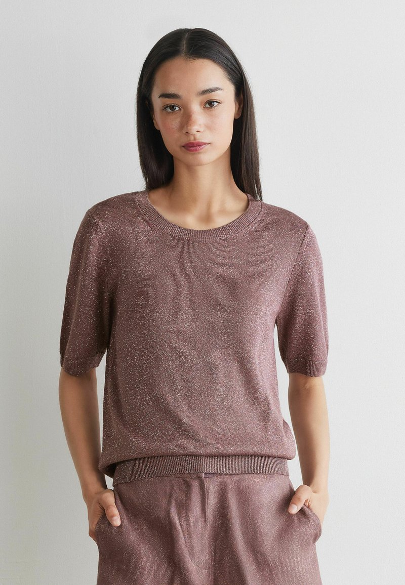 Jonge vrouw met lang donker haar, gekleed in een roze gebreide top met korte mouwen en bijpassende broek, staand tegen een effen lichte muur.