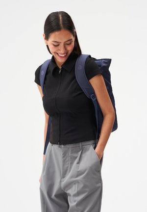 Lächelnde Frau mit langen dunklen Haaren trägt ein schwarzes kurzärmeliges Zip-up-Oberteil, graue Hose und einen marineblauen Rucksack, die Hände in den Taschen, schaut nach unten.