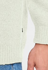 Suéter de punto verde claro con acabado texturizado y puños acanalados. Presenta una costura lateral con una pequeña etiqueta de logo cerca del dobladillo.