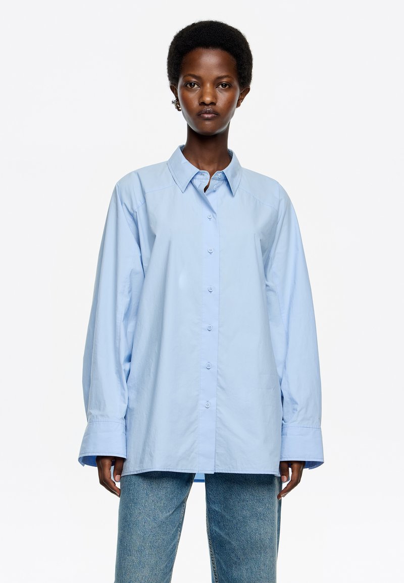 Lichtblauw button-up shirt met een kraag, lange mouwen en een losvallende pasvorm. Heeft een soepele textuur en een subtiele glans. Gecombineerd met blauwe jeans.