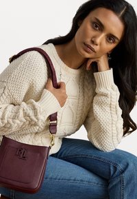 Burgundijas krāsas ādas kauss soma ar izspiesto logotipu, zelta toņu piederumiem un regulējamu siksnu; teksturēta adīta krēma krāsas džemperis.