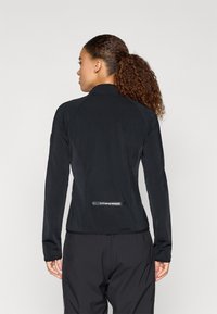 Juoda sportinė striukė su prigludusia forma, aukštu apykakle ir ilgomis rankovėmis. Yra logotipo detalė apatinėje nugaros dalyje ir glotni tekstūra.