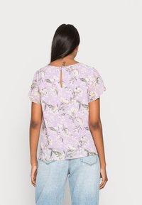 Blusa floral lilás com mangas curtas, apresentando um detalhe em forma de chave nas costas e um tecido suave, complementada por um padrão floral em tons suaves.