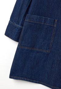 Denim jas in diepblauw met een grote voorzak. Heeft contrasterende oranje stiksels en een verlengd mouwontwerp. Gladde textuur.