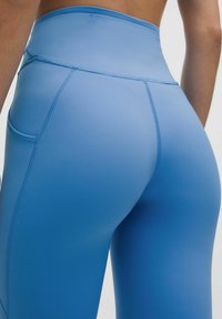 Zilas augstās jostas bikses, kas izgatavotas no gludas, elastīgas auduma. Aprīkotas ar sānu kabatām un kontrastējošu šuvju. Ideālas aktīvajai apģērbam.