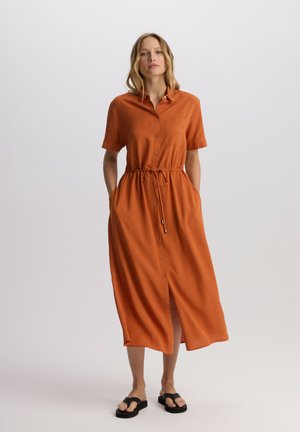 Długa sukienka z guzikami w pomarańczowym materiale, z krótkimi rękawami, paskiem w talii, kieszeniami po bokach i rozcięciem z przodu dla swobody ruchów.